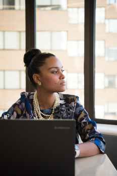 Photo of a young woman with a computer with her hair in a bun wearing pearls.