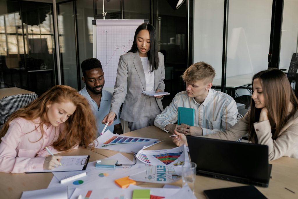 Image of a young Asian woman standing up pointing at a chart while her team of four other young professionals work or also look at the chart.