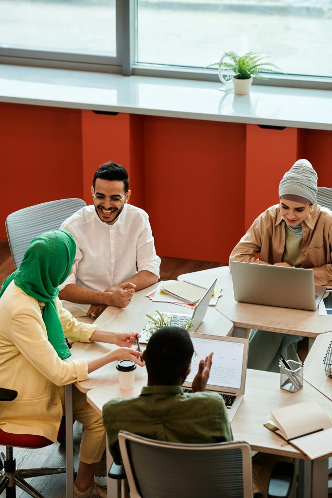 Image of a team of four sitting at a table. There are two men and two women. One man is Black and his back is to us. The two women are Muslim and wearing head scarfs.